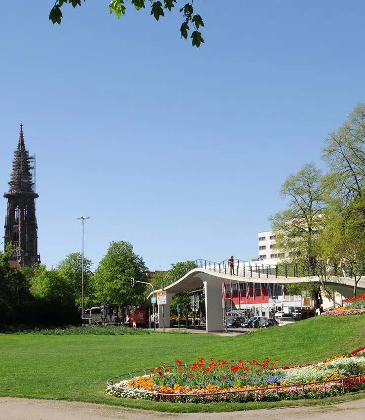 Eine sanft geschwunge Fußgängerbrücke, ein gut gepflegter Rasen und ein schickes buntes Blumenfeld im Vordergrund. Am linken Bildrand ist der Freiburger Münsterturm zu sehen. Der Himmel strahlend blau.